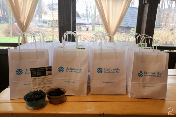 Papierowe torby z produktami z Uzdrowiska Goczałkowice-Zdrój ułożone obok siebie na stole. Z lewej strony ulotki i dwie miseczki z borowiną.