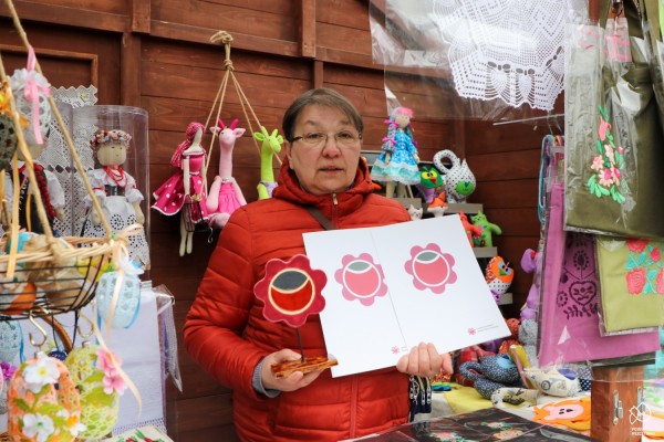 Pani Bożena Rokita stoi przed swoim stoiskiem. Trzyma w jednej ręce certyfikaty Lokalnej Marki Ziemi Pszczyńskiej, a w drugiej statuetkę przedstawiającą różę - logo marki.