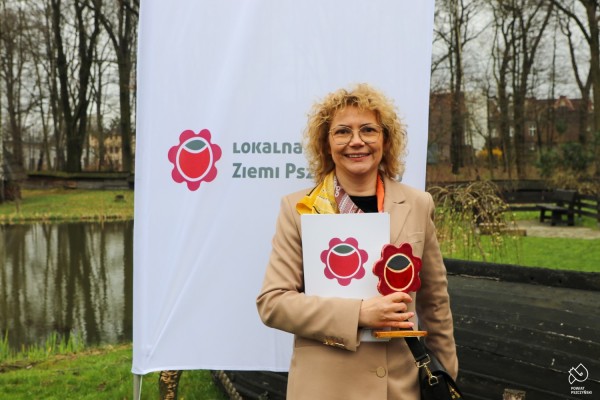 Przedstawicielka Karczmy Wiejskiej stoi na tle flagi z logo Lokalnej Marki Ziemi Pszczyńskiej i trzyma certyfikaty oraz statuetkę przedstawiającą różę - logo marki.