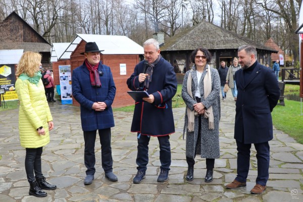 Prowadzący przemawia przez mikrofon, obok stoją przedstawiciele Powiatu Pszczyńskiego oraz Skansenu Zagroda Wsi Pszczyńskiej