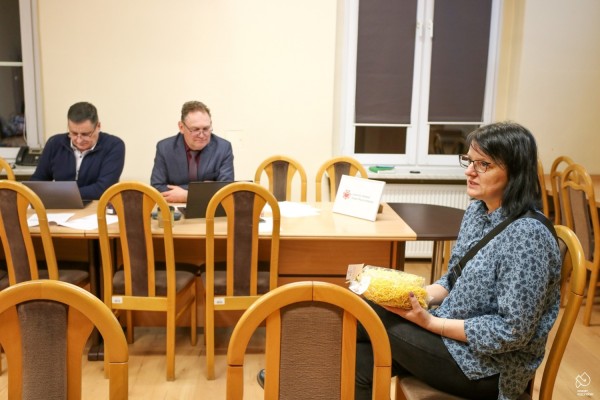 Kobieta trzymająca w ręku paczkę makaronu siedzi na krześle na środku sali. Przy stołach siedzi dwóch przedstawicieli Komisji Certyfikacyjnej, spoglądają w ekrany laptopów.