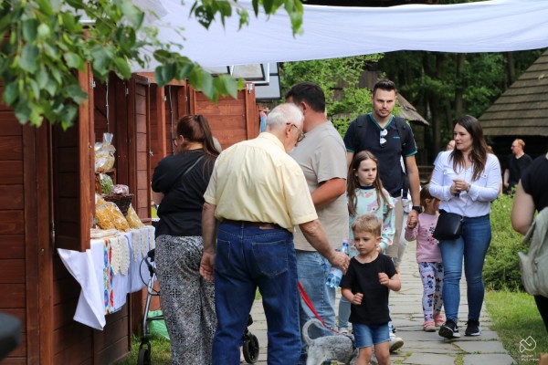 Ludzie spacerujący w skansenie obok drewnianych budek oraz oglądający produkty przy stoiskach.