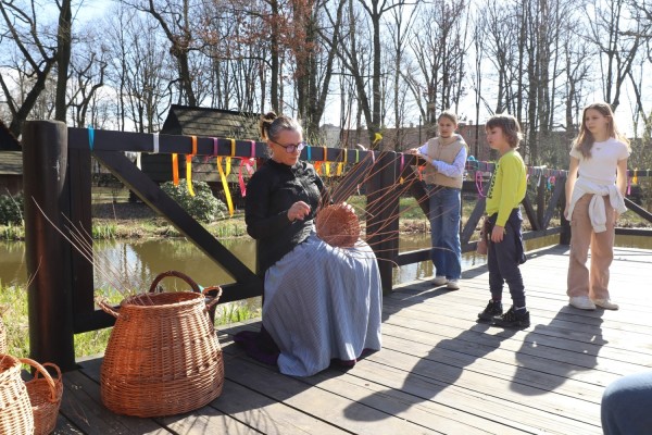 Na mostku siedzi na krzesle pani wyplatająca kosze z wikliny, obok przypatruje się jej trójka dzieci.