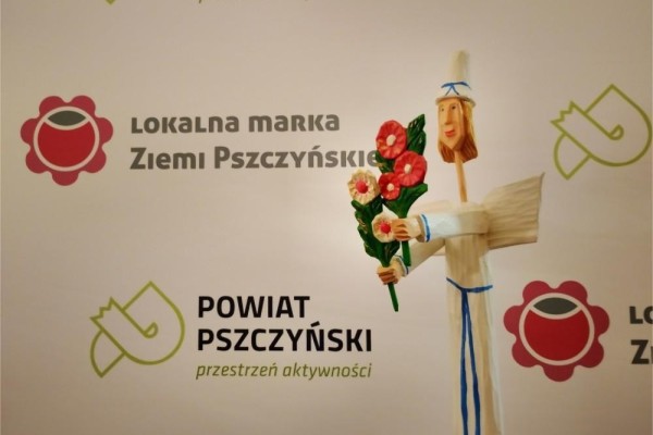 Drewniana rzeźba anioła, w tle ścianka z logo Lokalnej Marki Ziemi Pszczyńskiej oraz logo Powiatu Pszczyńskiego.