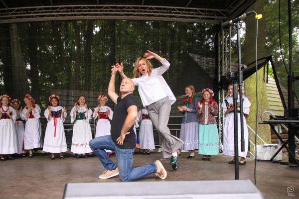 Na scenie tańczący prowadzący, za nimi stoi dziecięcy zespół folklorystyczny.